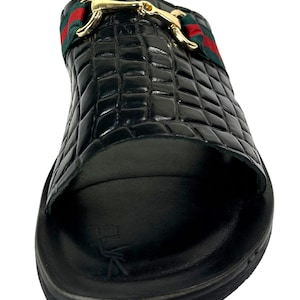 May include: A black slide sandal featuring a crocodile-embossed texture on the upper strap. The strap is accented with a green and red striped fabric band and a polished gold-tone metal horsebit detail. The footbed is black with a logo imprint.