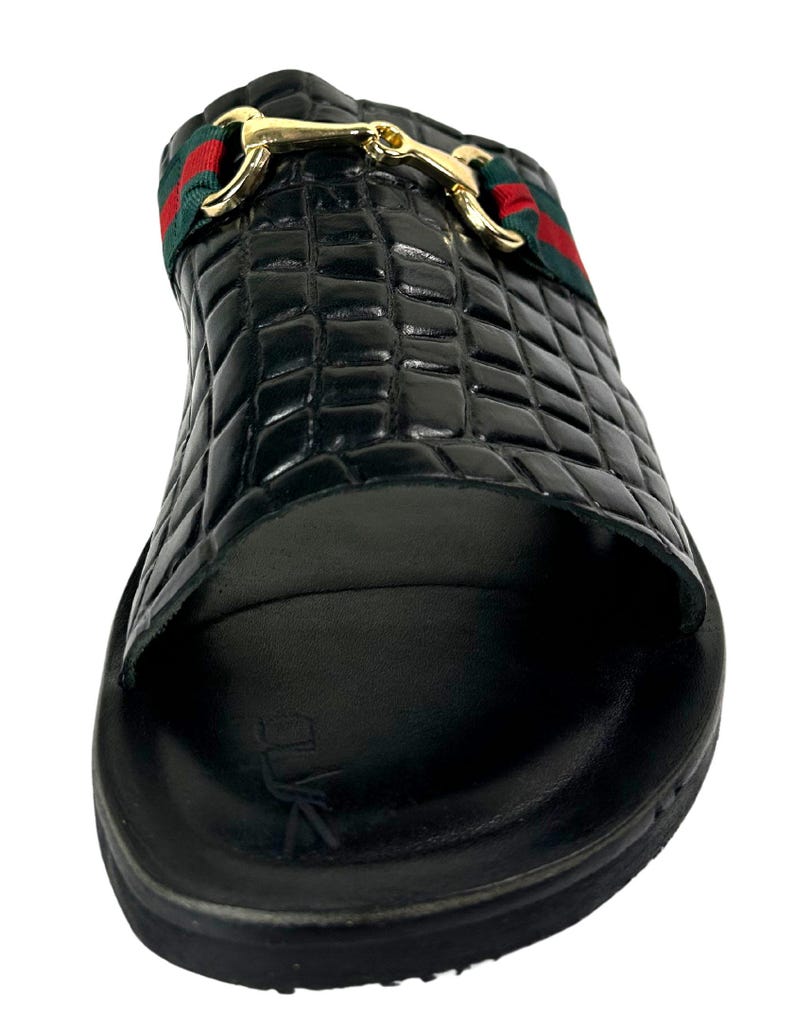 May include: A black slide sandal featuring a crocodile-embossed texture on the upper strap. The strap is accented with a green and red striped fabric band and a polished gold-tone metal horsebit detail. The footbed is black with a logo imprint.