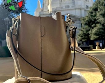 May include: A taupe leather bucket bag with a detachable, striped shoulder strap. The bag has silver-tone hardware and a small handle. The image includes a "Made in Italy" stamp.