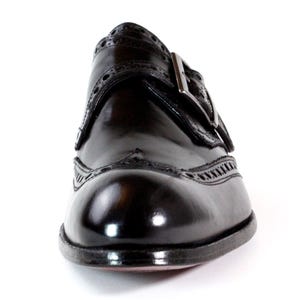 May include: A polished dark brown leather monk strap shoe featuring intricate brogue detailing along the toe cap and side panels. The shoe includes a single metallic buckle closure on the side and a rounded toe, showcasing a smooth finish.