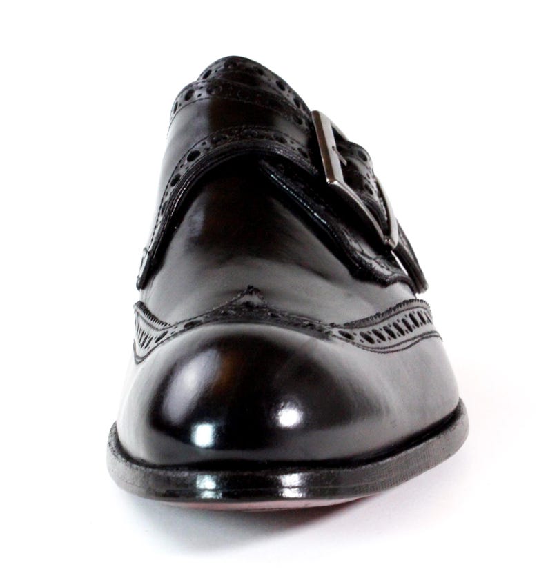 May include: A polished dark brown leather monk strap shoe featuring intricate brogue detailing along the toe cap and side panels. The shoe includes a single metallic buckle closure on the side and a rounded toe, showcasing a smooth finish.