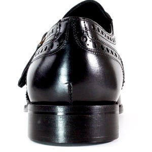 May include: A polished black leather dress shoe viewed from the back, featuring intricate brogue detailing along the heel counter and a stacked leather heel. The shoe has a smooth, glossy finish and a structured silhouette with visible stitching.