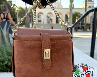 May include: A structured brown handbag featuring a combination of smooth leather and suede textures. The bag includes a gold-tone metal turn-lock closure on a vertical tab and a matching gold-tone chain shoulder strap for carrying.