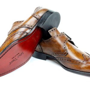 May include: A pair of tan leather monk strap shoes with brogue detailing and a wingtip toe. The shoes feature a double buckle closure, a black heel, and a smooth red sole with the text Ivar Troy Made in Italy embossed on the bottom surface.