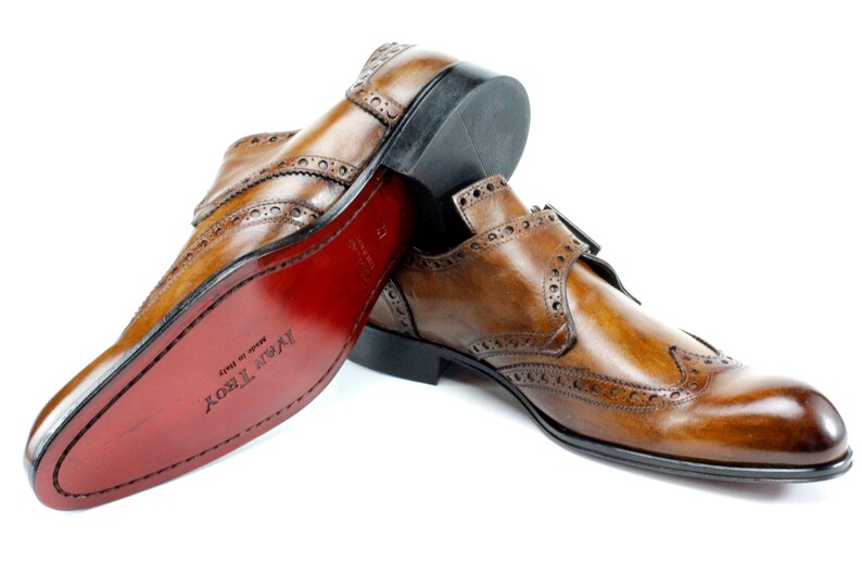 May include: A pair of tan leather monk strap shoes with brogue detailing and a wingtip toe. The shoes feature a double buckle closure, a black heel, and a smooth red sole with the text Ivar Troy Made in Italy embossed on the bottom surface.
