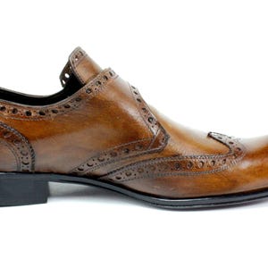 May include: A brown leather shoe with a wingtip design, featuring decorative brogue perforations along the seams and edges. The shoe has a smooth, polished finish, a rounded toe, and a solid black heel and sole, presented from a side profile.