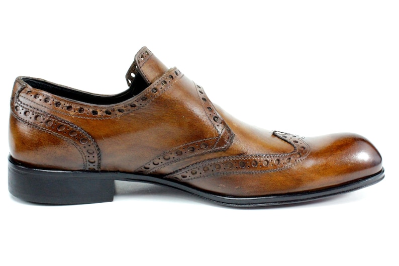 May include: A brown leather shoe with a wingtip design, featuring decorative brogue perforations along the seams and edges. The shoe has a smooth, polished finish, a rounded toe, and a solid black heel and sole, presented from a side profile.