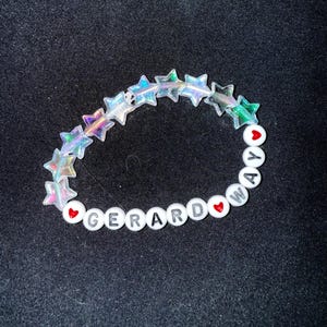 May include: A beaded bracelet with iridescent star-shaped beads and white round beads. The white beads spell out "GERARD WAY" with red heart beads separating the words. The bracelet is on a black background.