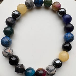 May include: A beaded bracelet featuring a variety of colorful, spherical gemstones. The beads include shades of blue, green, yellow, black, and gray, with some displaying unique patterns. The bracelet is arranged in a circular shape.