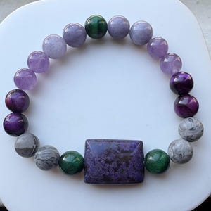 May include: A beaded bracelet featuring a variety of colorful stones. The bracelet includes round beads in shades of purple, green, and gray, along with a rectangular purple stone. The bracelet is displayed on a white surface.
