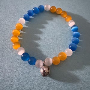 May include: A beaded bracelet featuring a baseball charm. The bracelet is made of blue, white, and orange beads. The baseball charm is silver-colored. The bracelet is on a light blue surface.