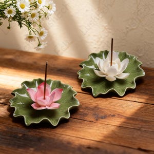 May include: Two ceramic incense holders shaped like lotus leaves, each with a lotus flower and an incense stick. One flower is pink, the other white. The holders are green and sit on a wooden surface.
