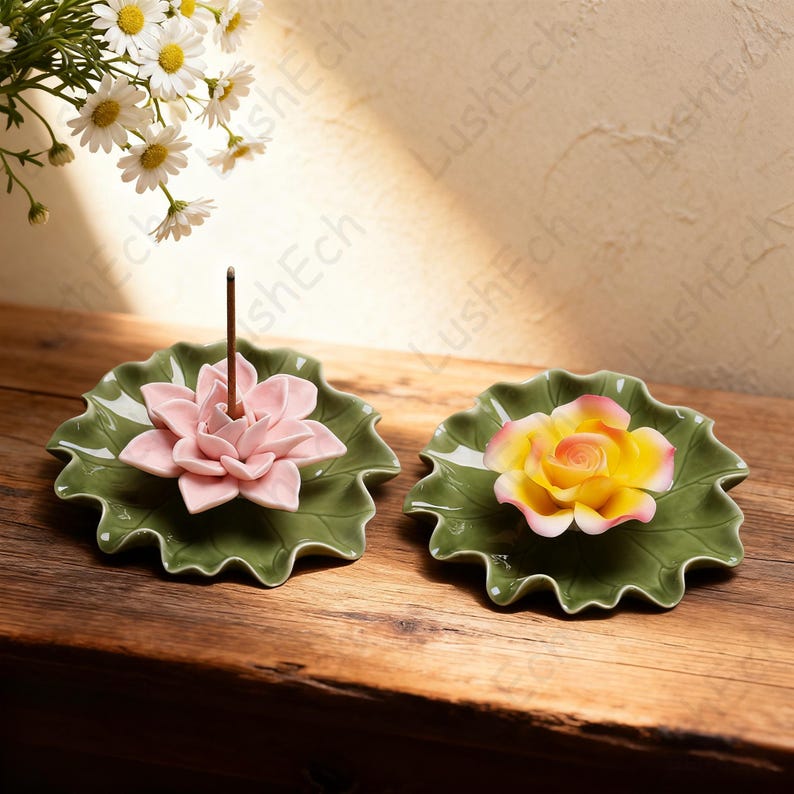 May include: Two ceramic incense holders shaped like lily pads, each with a flower. One has a pink lotus flower, the other a yellow rose. The holders are green and sit on a wooden surface.