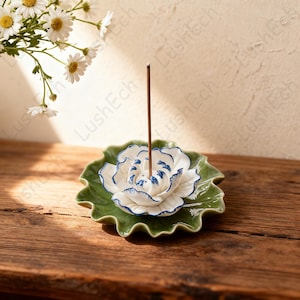 May include: A ceramic incense holder shaped like a lotus flower. The holder features a white flower with blue accents, set on a green leaf-shaped base. An incense stick stands upright in the center. The holder sits on a wooden surface.
