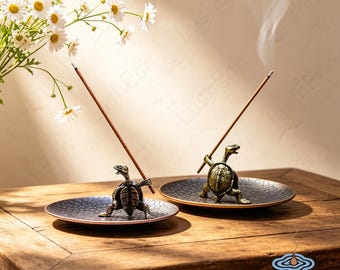 May include: Two bronze-colored turtle incense holders, each positioned on a circular tray with a textured, dark, scale-like pattern. Each turtle figure holds a single lit incense stick, with thin trails of smoke rising into the air.
