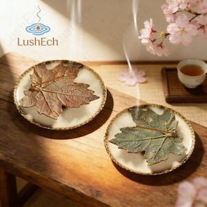 May include: Two ceramic plates with a cream base and brown rim, each with a detailed leaf design. One leaf is brown, the other green. The plates are on a wooden surface, with a cup of tea and pink flowers in the background.