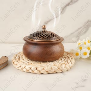 May include: A brown wooden incense burner with a bronze lid, releasing a wisp of smoke. The burner rests on a woven straw mat. White flowers and a small wooden object are in the background. The burner is a decorative item.
