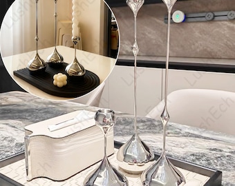 May include: A collection of silver candle holders with a teardrop-shaped base and long, slender stems. The candle holders are displayed on a table with a white tissue box and a black tray. The overall theme is home decor.