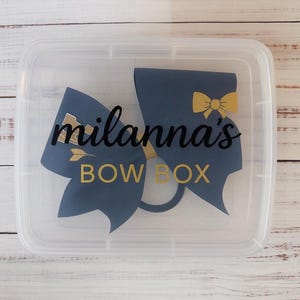 May include: A clear plastic box featuring a navy blue bow design with gold detailing. The text "milanna's BOW BOX" is printed on the box. The box is designed for storing hair bows.