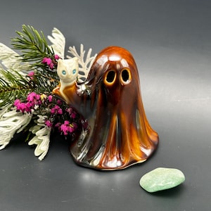 Puede incluir: Una decoración de Halloween de cerámica marrón brillante con un fantasma con dos agujeros para los ojos y un pequeño gato blanco con ojos azules. Una piedra verde y ramitas con vegetación y flores rosas están en el fondo.