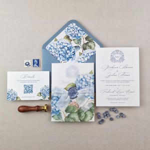 May include: Elegant wedding invitation suite featuring blue hydrangea floral designs. Includes invitation cards, an envelope, a details card, and a wax seal. The invitation cards have the names "Makara Barass & Joshua Acass" and the date "ON 10TH JUNE 2017".