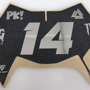 May include: Black and tan motorcycle number plate with the number 14 in silver. The plate has the text "PK!", "TG BUILT", "VALOR GRAFIX", and a "Monster" logo.
