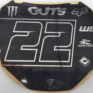 May include: Black and silver motocross number plate with the number 22 in white. The plate features the "GUTS" and "FOX" logos, along with other brand logos. The plate has a textured surface.