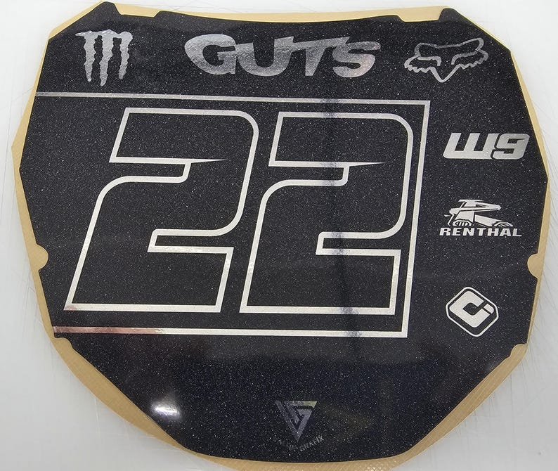 May include: Black and silver motocross number plate with the number 22 in white. The plate features the "GUTS" and "FOX" logos, along with other brand logos. The plate has a textured surface.