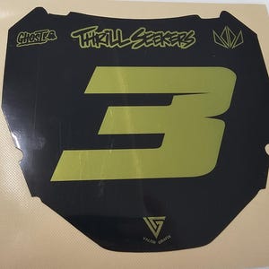 May include: Black and gold motocross number plate with the number 3. The plate features the text "Ghosts" and "Thrill Seekers" in a stylized font. The bottom of the plate has the text "Valor Grafix".
