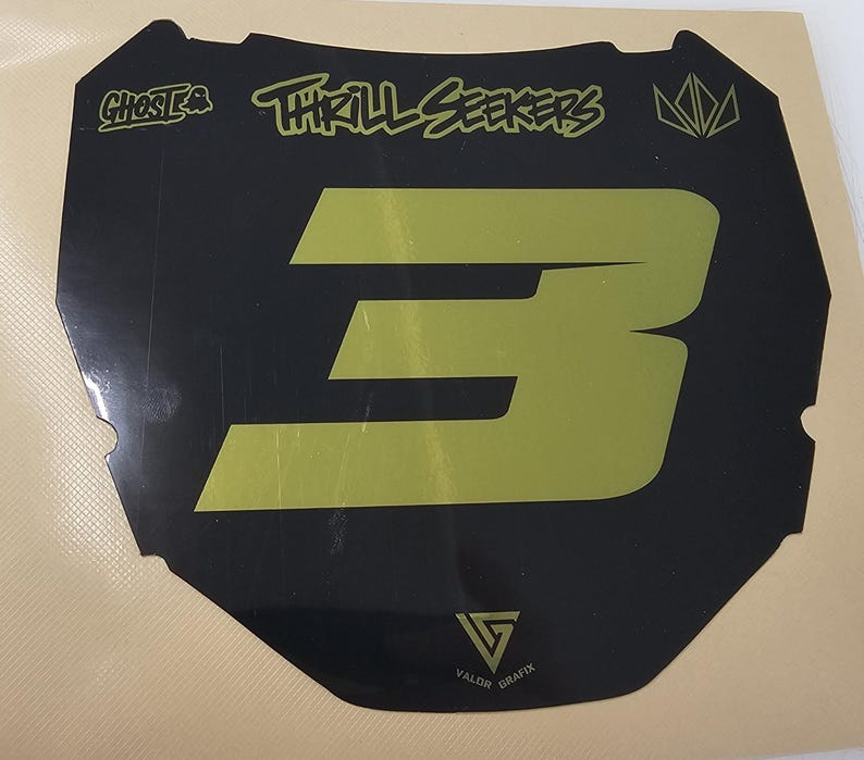 May include: Black and gold motocross number plate with the number 3. The plate features the text "Ghosts" and "Thrill Seekers" in a stylized font. The bottom of the plate has the text "Valor Grafix".