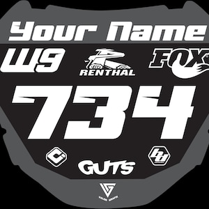 May include: A black and gray motocross number plate with the text "Your Name" at the top. The number "734" is prominently displayed in white, along with the logos for FOX, RENTHAL, and GUTS. Additional logos and the letters "W9" are also visible.