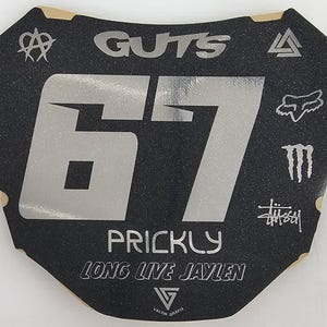 May include: Black and silver motocross number plate with the number 67. The plate features the word "GUTS" at the top, the name "PRICKLY" and "LONG LIVE JAYLEN" at the bottom, and various logos.