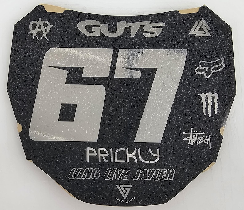 May include: Black and silver motocross number plate with the number 67. The plate features the word "GUTS" at the top, the name "PRICKLY" and "LONG LIVE JAYLEN" at the bottom, and various logos.