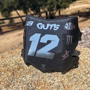 May include: Black and silver motocross number plate with the number 12 in large silver font. The plate has the text "19 GUTS" and various logos. The plate is sitting on a rock.