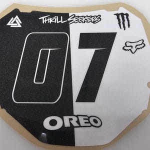 May include: A black and white motocross number plate with the number "07" in a large font. The plate features the text "THRILL SEEKERS" and the word "OREO". Logos for Monster Energy and Fox Racing are also visible.