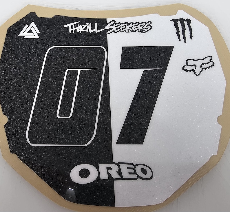 May include: A black and white motocross number plate with the number "07" in a large font. The plate features the text "THRILL SEEKERS" and the word "OREO". Logos for Monster Energy and Fox Racing are also visible.