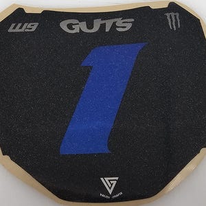 May include: Black and blue number plate with the number 1 in blue. The word "GUTS" is in silver at the top, with "W9" on the left and a Monster Energy logo on the right. The bottom has the Valor Grafix logo.