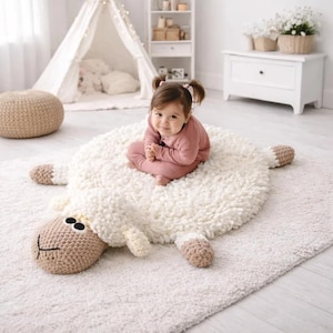 Puede incluir: Una alfombra blanca con forma de oveja con cabeza y patas de ganchillo. La alfombra tiene un aspecto suave y texturizado. La cabeza de la oveja es de color marrón claro con ojos negros. Un niño con ropa rosa está sentado en la alfombra.