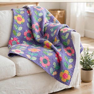 May include: A purple quilt featuring a floral pattern with yellow, pink, and green flowers and stems. The text at the top reads Flower Meadow Quilt Pattern 157.48 cm x 203.2 cm • PDF Download. The quilt is draped over a white sofa.