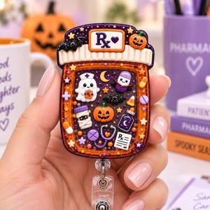 May include: A Halloween-themed badge reel shaped like a prescription bottle with a purple and orange color scheme. The bottle features a white cap with the text Rx and a heart, decorated with small charms including a ghost, pumpkin, bat, and stethoscope.