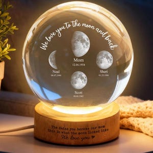 May include: A clear crystal ball with phases of the moon and names and dates etched inside. The ball sits on a wooden base that is lit from within. Text on the ball reads "We love you to the moon and back". The base has text that reads "The dates you became our mom this is what the moon looked like We love you".