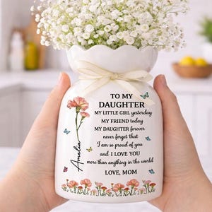 May include: White ceramic vase with a floral arrangement and a cream ribbon. The vase features a floral design, butterflies, and the text "TO MY DAUGHTER" and a personalized message. The vase is held by a person.
