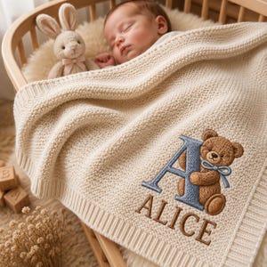 May include: A cream-colored knitted baby blanket featuring an embroidered design of a blue letter A, a brown teddy bear with a blue bow, and the name ALICE in brown lettering. The blanket has a ribbed border and a textured knit pattern throughout.