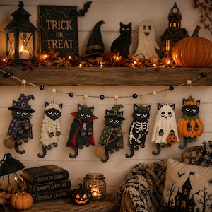 May include: A Halloween garland featuring seven hanging black cat ornaments dressed in various costumes, including a witch, mummy, vampire, wizard, skeleton, ghost, and pumpkin. The cats are suspended from a string with black and white beads.