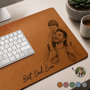 May include: A brown leather desk mat with a black engraved image of a father carrying his son on his shoulders. The text "Best Dad Ever" is written in script. A white keyboard is on the left.
