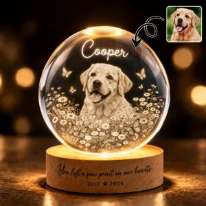 Puede incluir: Una lámpara de noche personalizada con forma de bola de cristal con la imagen de un perro, el nombre "Cooper", mariposas y flores. La base de madera tiene la inscripción "You left a paw print on our hearts" y los años 2017 y 2026.