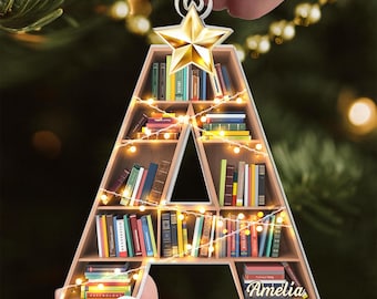 May include: A wooden Christmas decoration in the shape of the letter "A", filled with miniature books and fairy lights. A gold star sits at the top, and the name "Amelia" is printed at the bottom. The ornament hangs from a gold string.