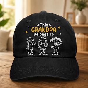 May include: Black baseball cap with the text "This Grandpa Belongs to" and the names Billie, Victoria, and Amelia. The text and illustrations are in white and yellow. The cap has a curved brim.