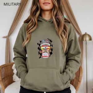 May include: Olive green hoodie with a graphic of a can wearing sunglasses, a mustache, and a hat. The can has the words "Raise Hell Praise" on the hat and "Merica" on the can. A black and white checkered flag is behind the can.