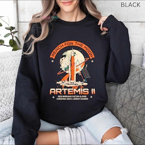 May include: Black sweatshirt with a graphic featuring a rocket launching towards the moon, with the text "REACH FOR THE MOON" and "ARTEMIS II". The names of the crew members are listed below the text. The sweatshirt is a crew neck style.